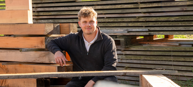 Person sitzt auf Holzbalken und blickt in die Kamera