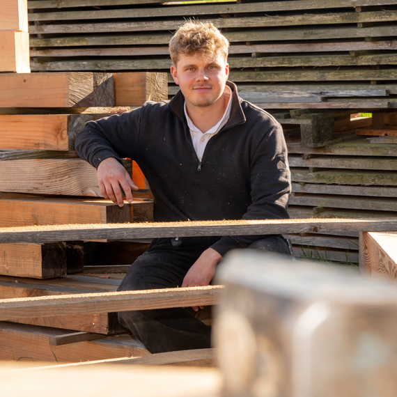 Person sitzt auf Holzbalken und blickt in die Kamera